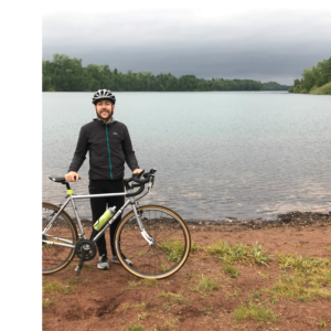 A person who presents masculine with a light skin tone stands on a beach of a lake with a bike and is wearing black clothing and a bike helmet.