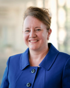 A person with an updo of light brown hair and light skin tone wears royal blue clothing with large buttons.