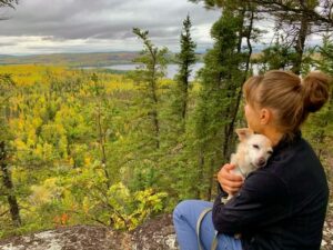 Kelley holds a small chihuahua mix while overlooking trees and Lake Superior from a cliff.