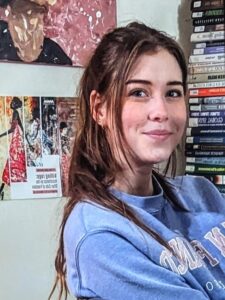 Ava is a younger adult with long brown-red hair, brown eyes, and light skin. Ava wears a sky blue sweatshirt and is posed near a tall stack of books in a room with art and educational posters on the walls.