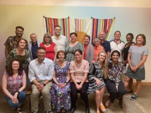 Members of HPHC's board, staff, and fellowship program gather at a celebration dinner and sit and stand in rows in front of a colorful tapestry on the wall behind them.