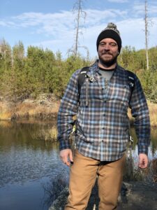 Alec, a young to middle-aged person with light skin tone and brown facial hair is outside on a hike with a backpack and outdoor gear.