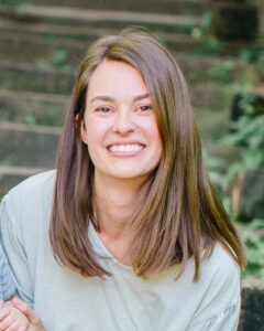 Brenna, a young to middle-aged person with light skin tone and straight, shoulder-length light brown hair wears a cream top. 