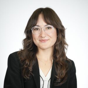 Hannah, a young person with light skin ton and feathery bangs and long dark brown-black hair with clear acetate glasses wears a black blazer and white top. 