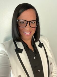 Myriam, a young to middle-aged adult Black woman with medium skin tone and short, straight black hair wears a white blazer and black glasses.